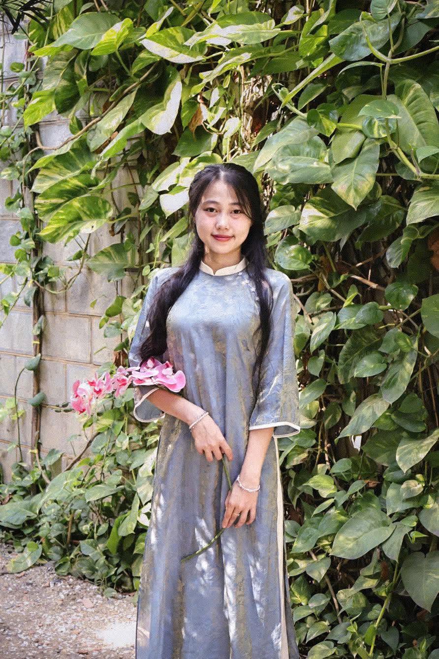 Khách mặc áo dài lụa xanh xám, cầm cành lan hồng bên giàn lá
